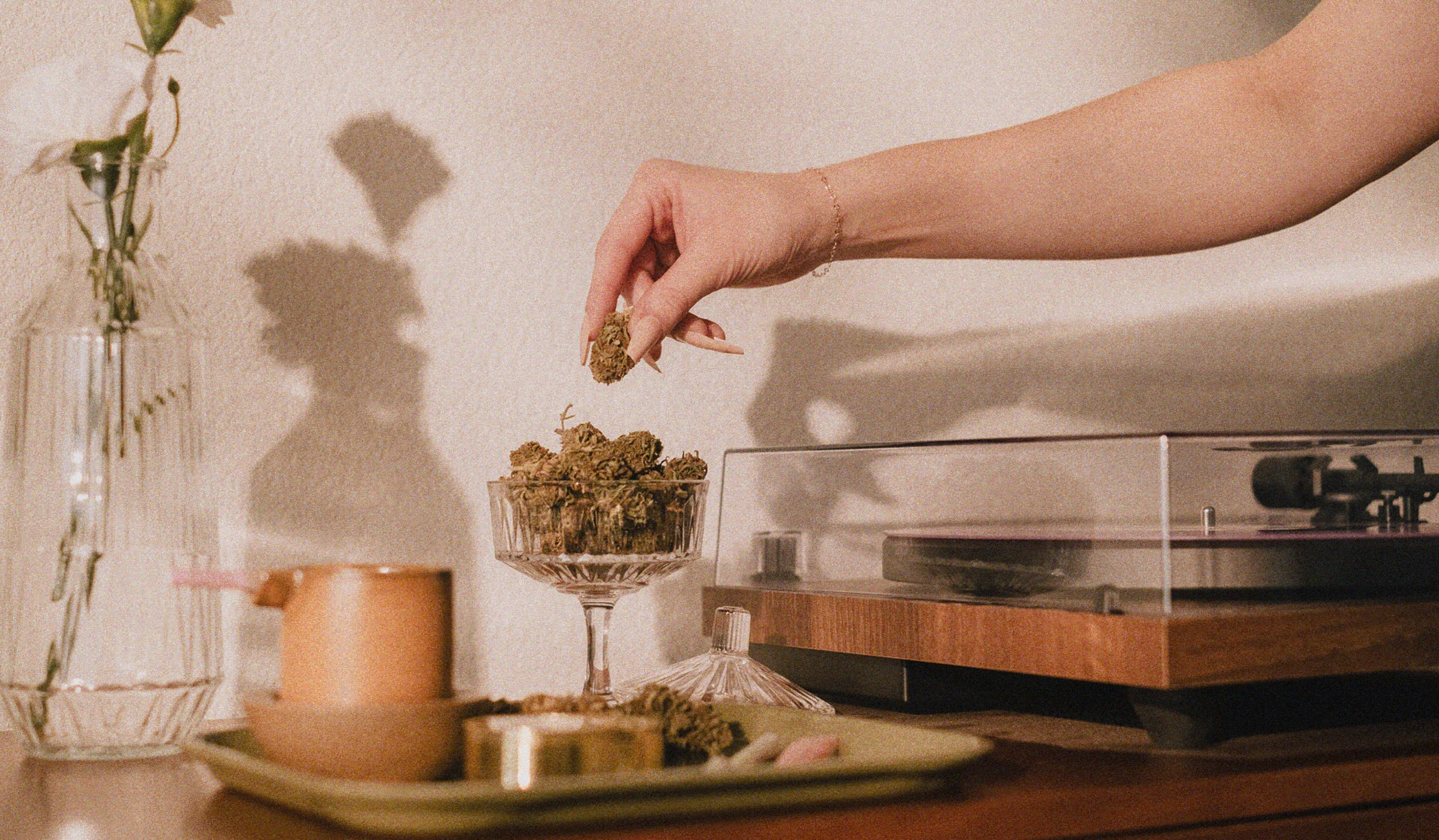 Harvesting tranquility: A woman's hand delicately picking a cannabis bud from an exquisite crystal cup in a cozy mid-century modern room adorned with a jar of flowers and a vintage vinyl record player.