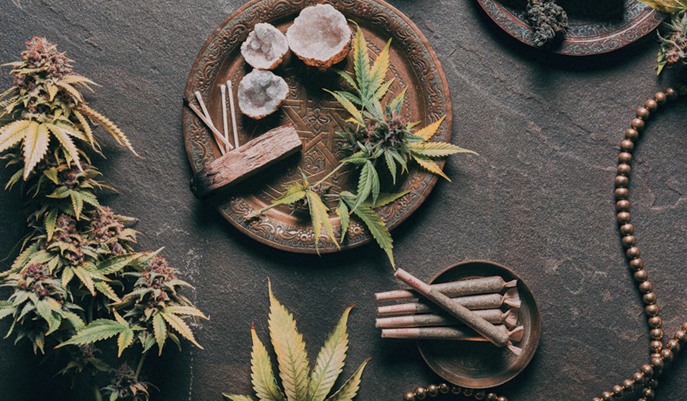 Elevated essentials: A captivating flatlay featuring plates on a dark table adorned with crystals, sage, pre-rolls, and a display of cannabis flowers and leaves.